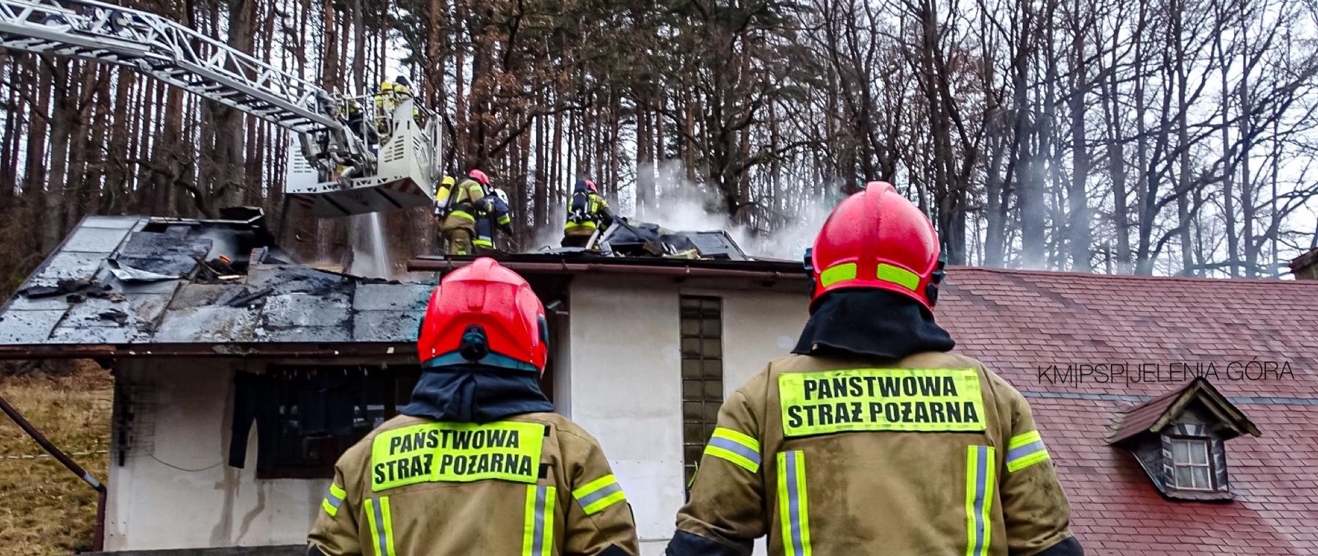 Na zdjęciu widać dwóch strażaków patrzących w kierunku budynku. Na dachu budynku widać strażaków oraz ulatniający się dym. Zdjęcie wykonane w dzień. Strażacy są w musztardowych mundurach. 