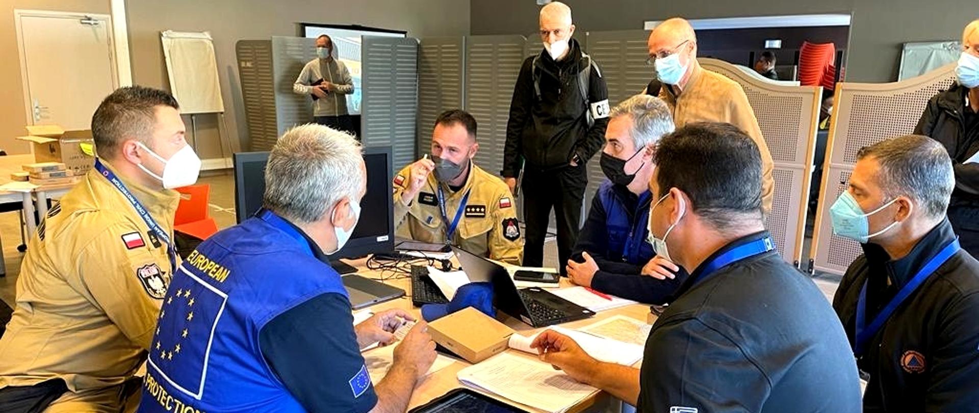 Dziesięciu mężczyzn w pomieszczeniu dydaktycznym. Sześciu z nich siedzi przy stoliku, pozostali stoją obok stolika. Wszyscy uczestnicy spotkania są w maseczkach ochronnych. 