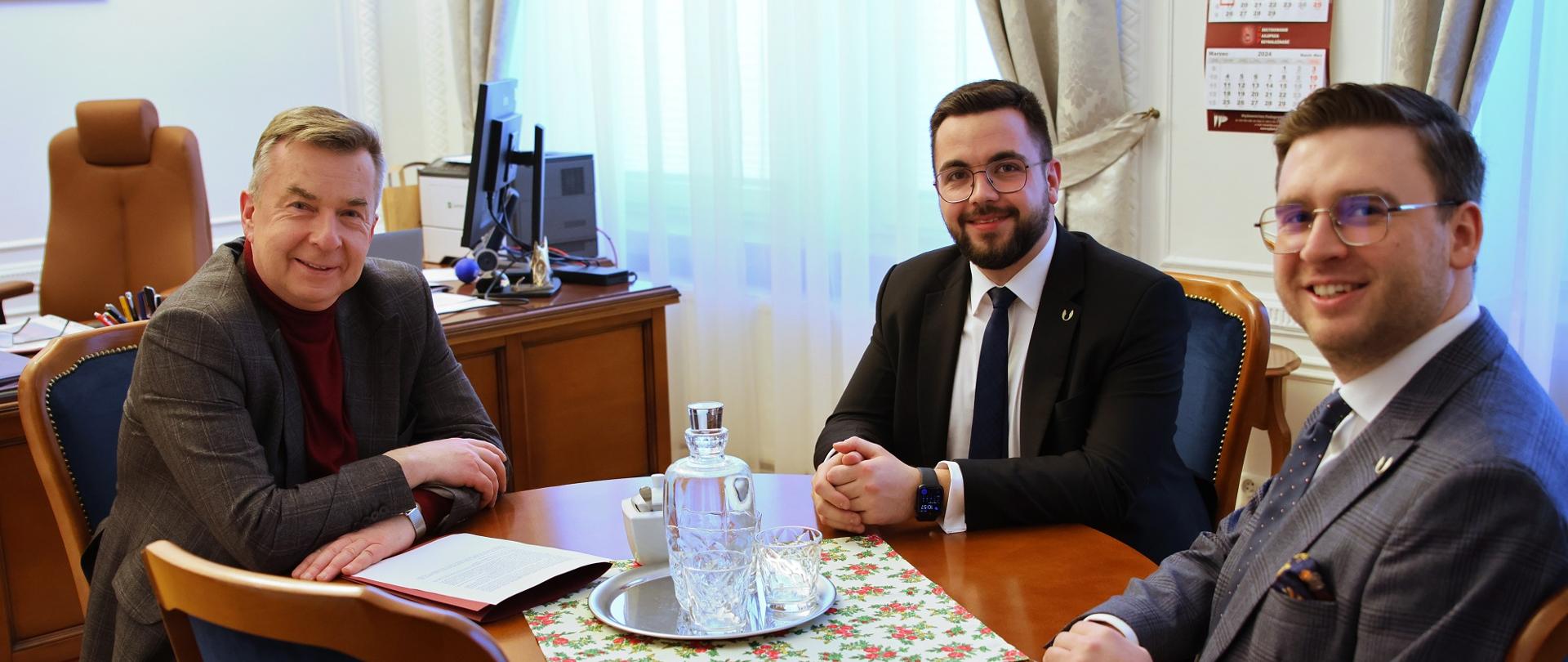 Przy drewnianym okrągłym stoliku siedzi minister Wieczorek i dwóch uśmiechniętych mężczyzn w garniturach, w tle zasłonięte firankami okno.