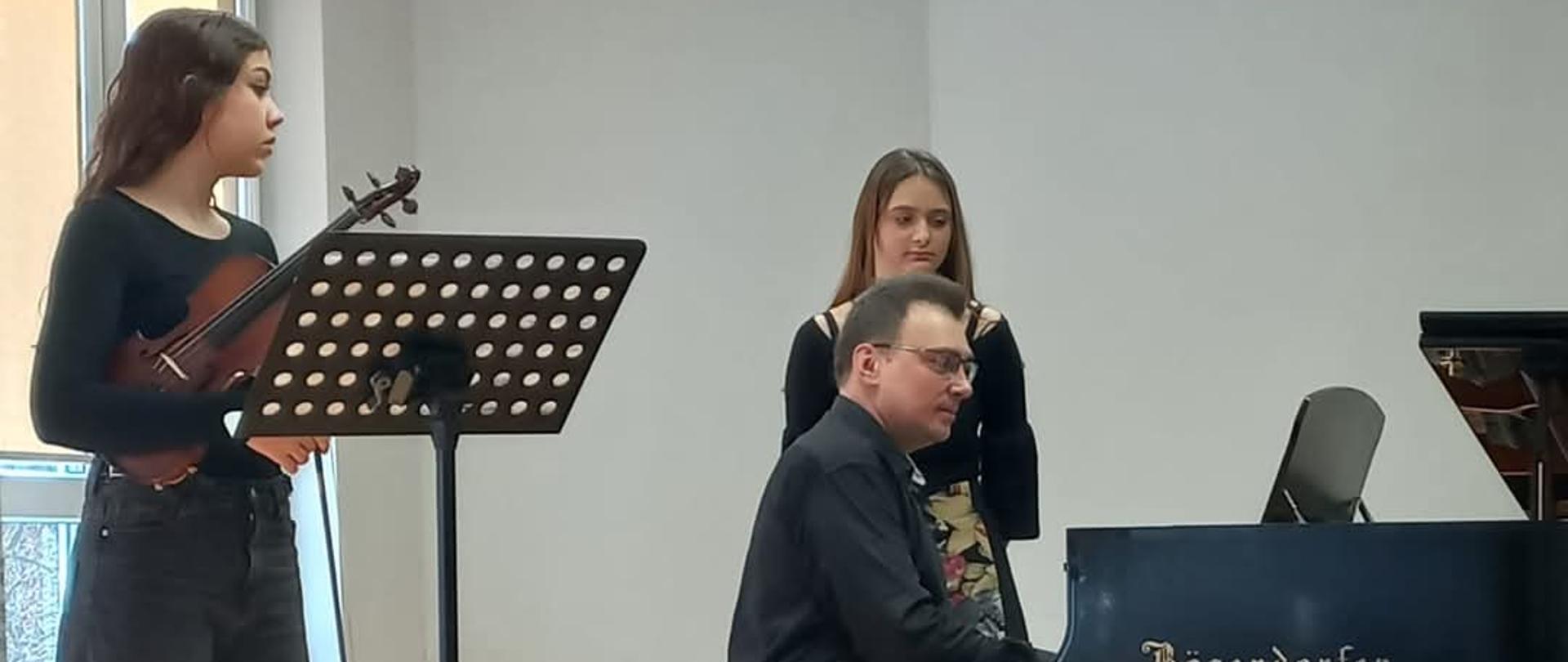 na zdjęciu meżczyzna grający na fortepianie obok niego stoją dwie dziewczyny jedna trzyma skrzypce