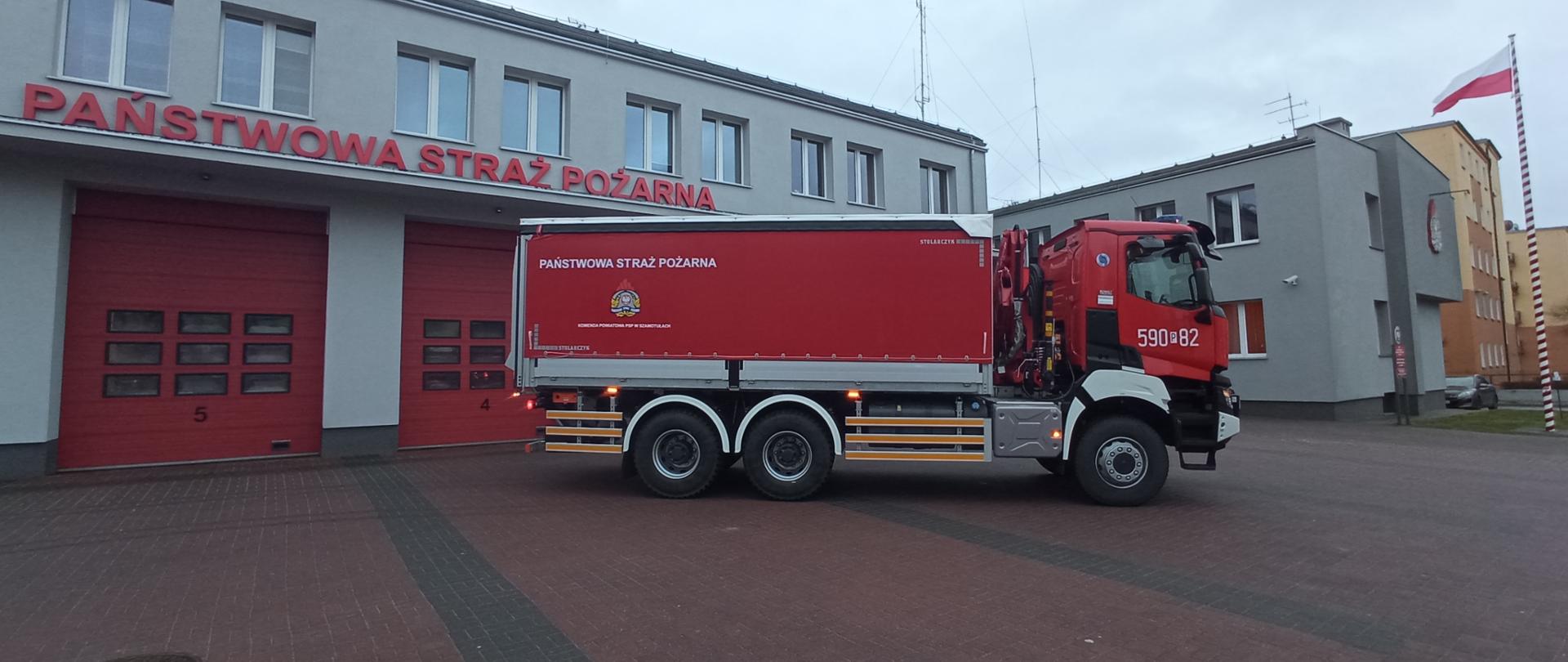 Ciężarowy samochód strażacki posiadający trzy osie jezdne, kabinę oraz zabudowę ładunkową znajdującą się za kabiną. Widok pojazdu z boku, z lewej strony. W zabudowie pojazdu znajduje się żuraw hydrauliczny oraz winda.