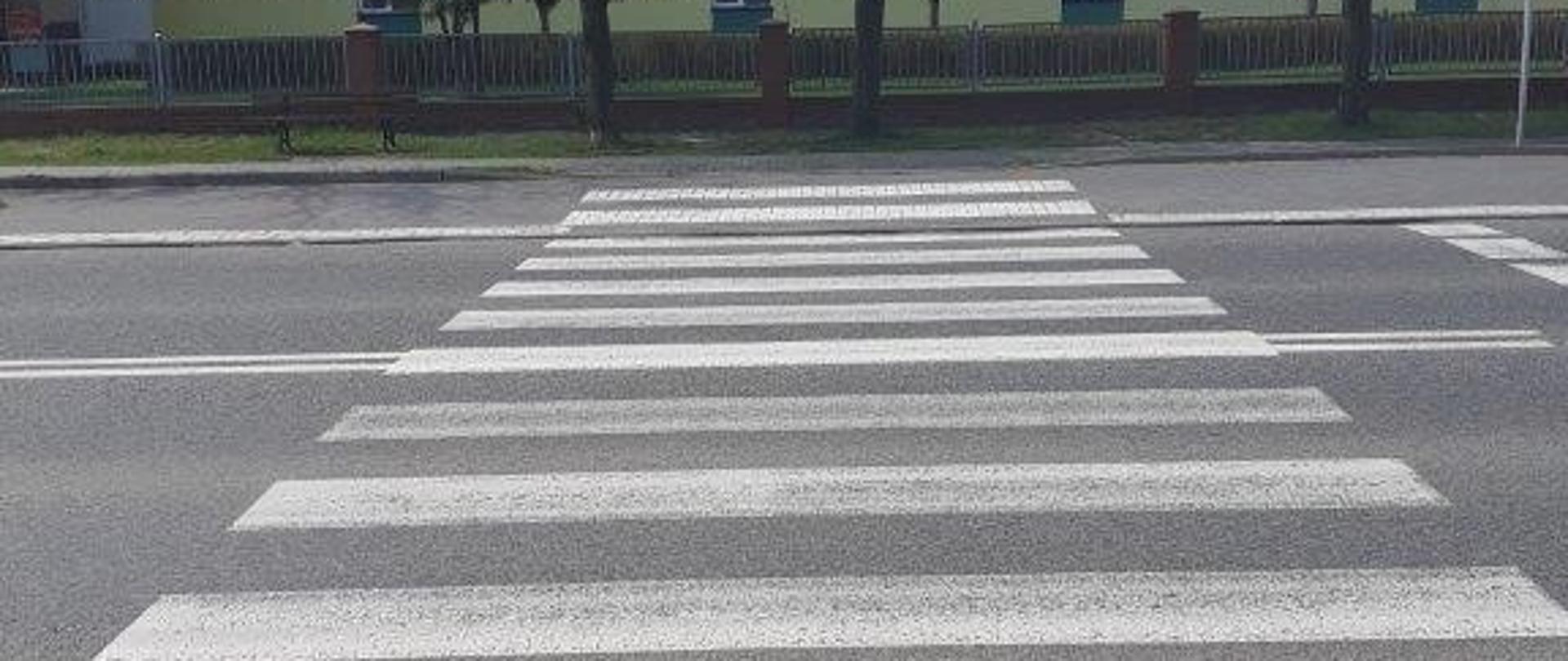 Zdjęcie przedstawia przejście dla pieszych przez drogę krajową w tle widoczny budynek piętrowy. 