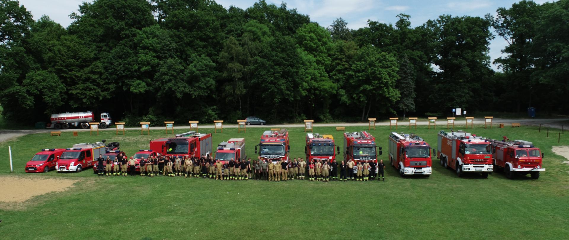 Strażacy i samochody strażackie stoją w szeregu na polanie.