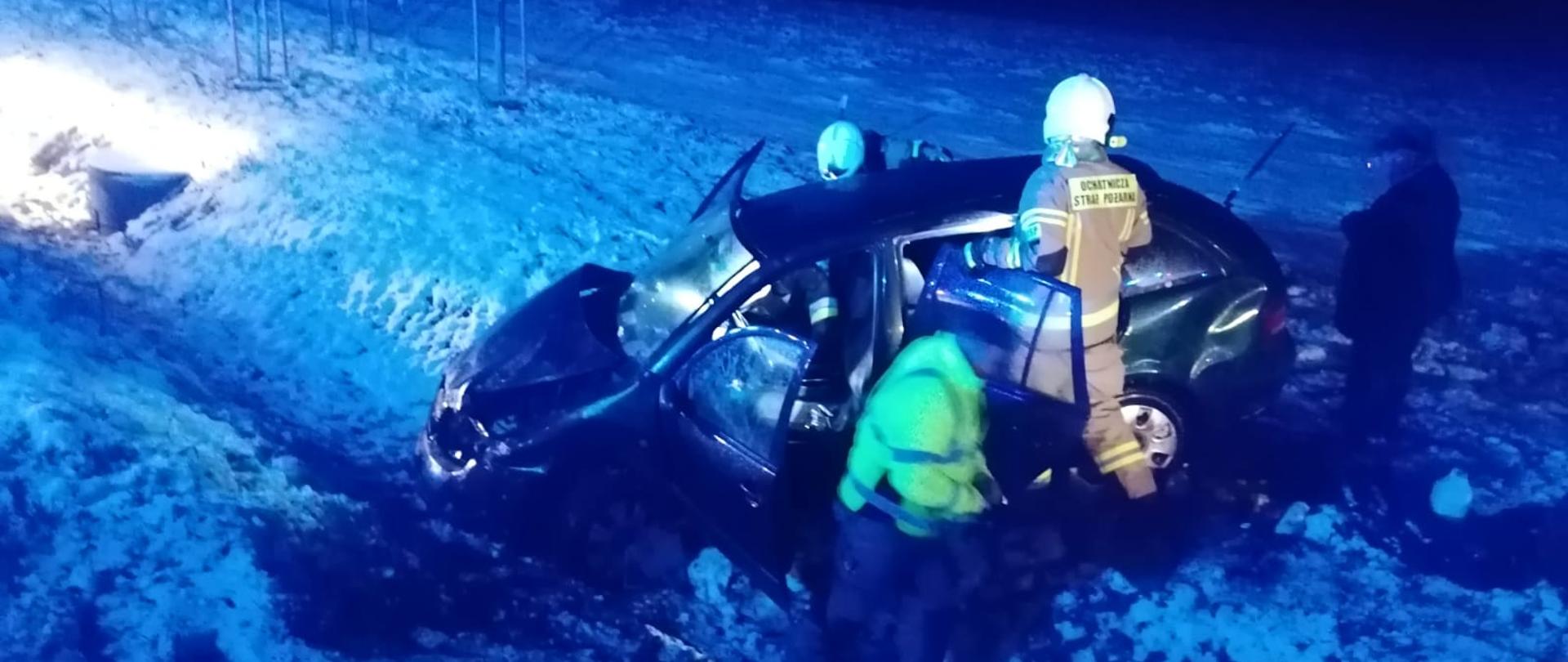Zdjęcie zrobione nocą. Dużo śniegu, biało, W rowie przy drodze stoi rozbite ciemne auto. Przy nim strażacy.