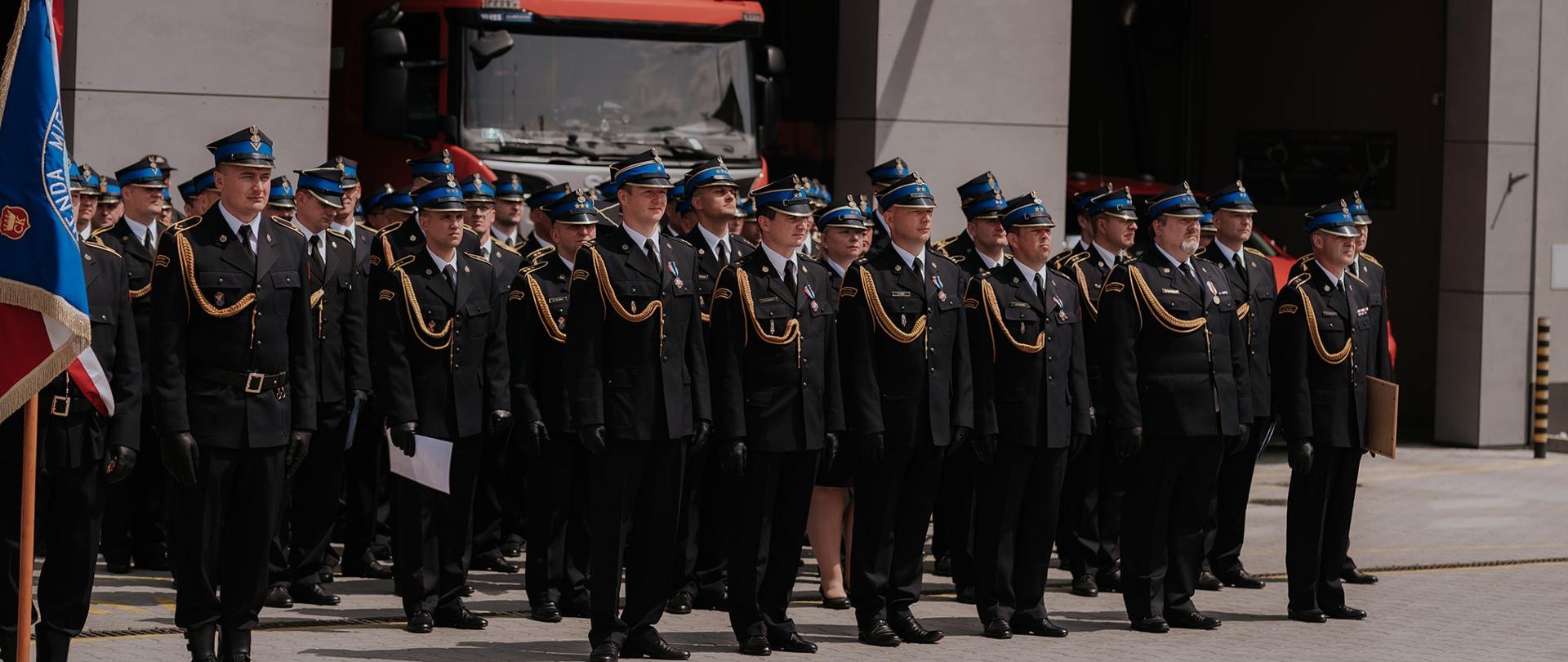 Przed budynkiem Jednostki Ratowniczo-Gaśniczej Numer 1 w Kielcach z otwartymi bramami, na tle zaparkowanych wewnątrz samochodów stoją ustawieni funkcjonariusze odznaczeni i awansowani.