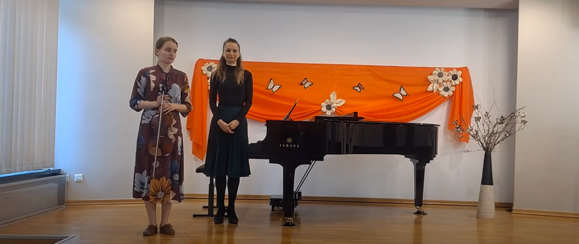 Na zdjęciu dwie osoby. Uczennica w brązowej sukience w kwiaty, trzyma skrzypce. Za nią na czarno ubrana akompaniatorka. Za nimi w tle czarny fortepian oraz dekoracja w kolorze pomarańczowym w z jasnymi motylkami i kwiatkami.