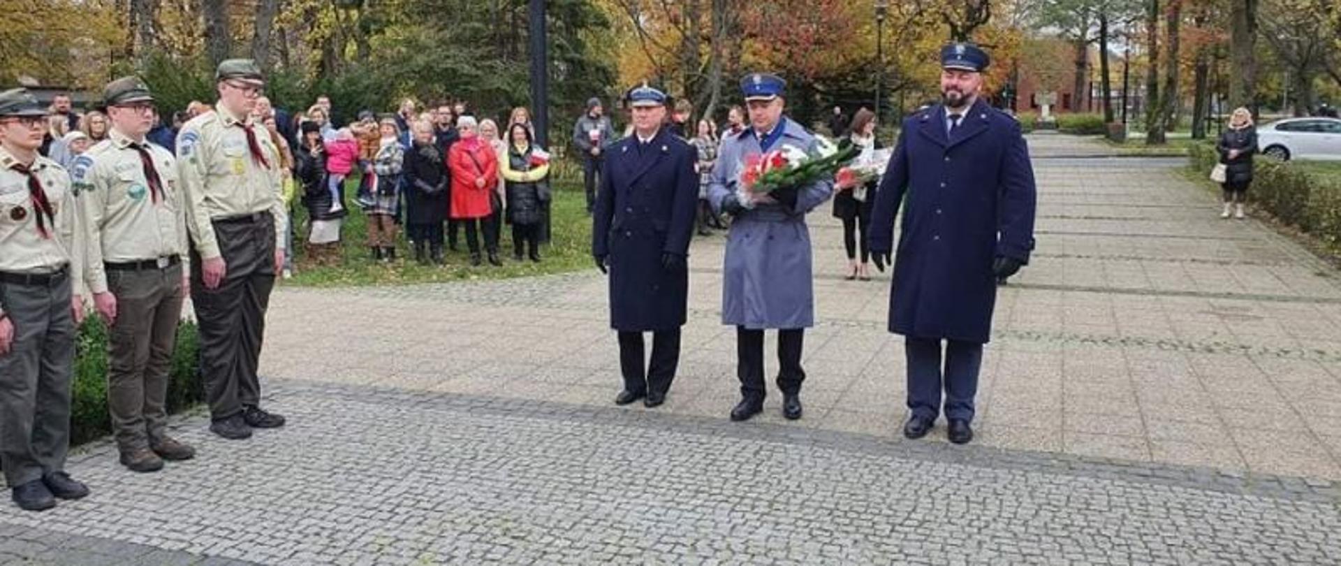 Na zdjęciu przedstawiciele policji, służb mundurowych i straży pożarnej.Wszyscy w mundurach galowych, maszerują z kwiatami w kierunku pomnika orła białego.