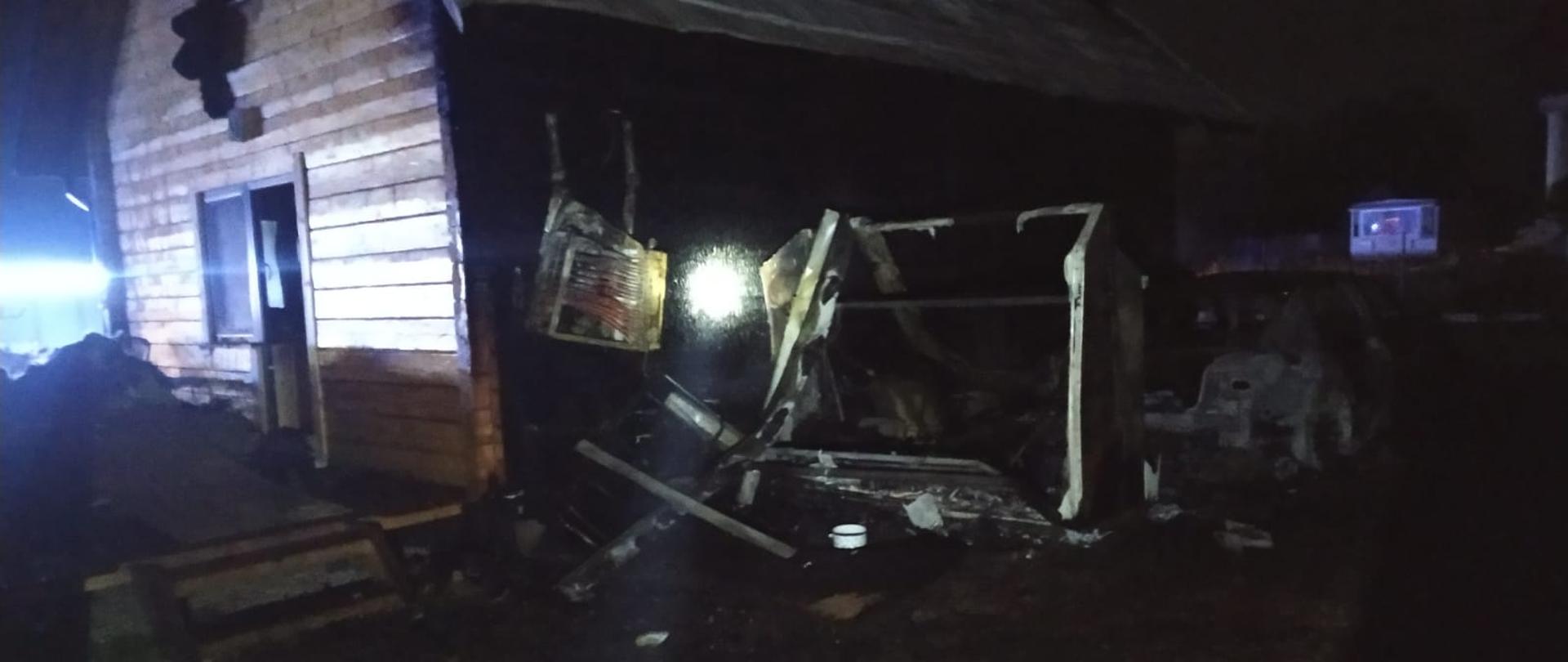 Zdjęcie przedstawia spaloną konstrukcję drewnianą domu letniskowego. Zdjęcie zrobione w porze nocnej. Po prawej stronie widnieje także spalona konstrukcja pojazdu osobowego.