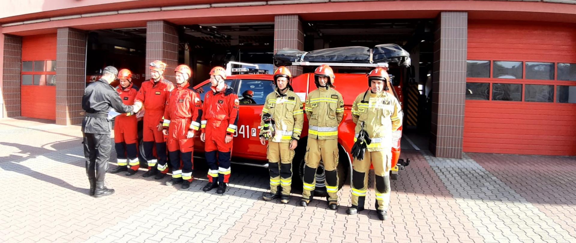 Zdjęcie przedstawia czterech strażaków ubranych w czerwone kombinezony ratownicze, na głowie czerwone kaski którzy odbierają nagrody od zastępcy dowódcy JRG1 w Kaliszu. Dowódca obrócony plecami do kadru, ubrany w mundur strażacki koloru czarnego, czapka strażacka na głowie,, na brzuchu zaciśnięty pas strażacki. Obok strażaków odbierających nagrody widzimy trzech strażaków stojących w rzędzie, mających na sobie ubranie specjalistyczne koloru piaskowego, na głowach czerwone chełmy z cyfrą jeden na przodzie. W tle widzimy samochód strażacki marki Ford, koloru czerwonego,. Na dachu samochodu czarny pokrowiec na sprzęt specjalistyczny. Dalej w tle widzimy otwarte wrota wyjazdowe JRG1 oraz część napisu " Państwowa Straż Pożarna JRG-1".. 