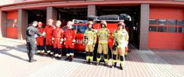 Zdjęcie przedstawia czterech strażaków ubranych w czerwone kombinezony ratownicze, na głowie czerwone kaski którzy odbierają nagrody od zastępcy dowódcy JRG1 w Kaliszu. Dowódca obrócony plecami do kadru, ubrany w mundur strażacki koloru czarnego, czapka strażacka na głowie,, na brzuchu zaciśnięty pas strażacki. Obok strażaków odbierających nagrody widzimy trzech strażaków stojących w rzędzie, mających na sobie ubranie specjalistyczne koloru piaskowego, na głowach czerwone chełmy z cyfrą jeden na przodzie. W tle widzimy samochód strażacki marki Ford, koloru czerwonego,. Na dachu samochodu czarny pokrowiec na sprzęt specjalistyczny. Dalej w tle widzimy otwarte wrota wyjazdowe JRG1 oraz część napisu " Państwowa Straż Pożarna JRG-1".