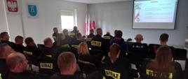 Na sali siedzi grupa kobiet i mężczyzn ubranych w mundury straży pożarnej patrząc na ekran z prezentacją
