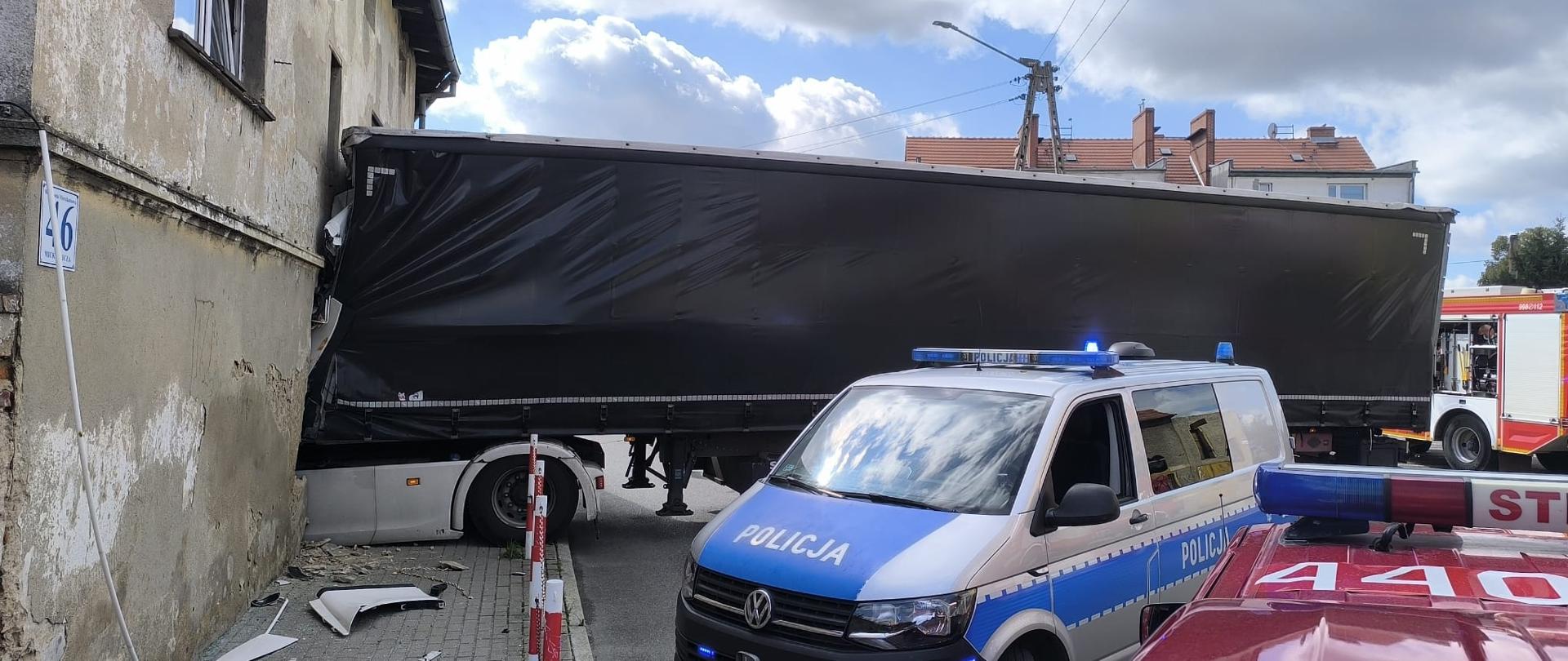 Ciężarówka uderzyła w ścianę budynku, z kabiną wbitą w ścianę. Obok stoją pojazdy służb – radiowóz policji i wóz strażacki – zabezpieczające miejsce zdarzenia.