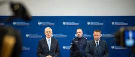 Minister Tomasz Siemoniak (po lewej), minister Marcin Kierwiński (po prawej) oraz komendant główny Policji gen. insp. Marek Boroń (w środku) stoją na tle niebieskiej ścianki ze zmuliplikowanym logo MSWiA (orzeł, napis Ministerstwo Spraw Wewnętrznych i Administracji oraz biało-czerwony pasek). Ministrowie stoją za szarymi mównicami z czarnymi mikrofonami kierunkowymi. Po prawej i lewej stronie zdjęcia widać rozmazane kamery telewizyjne. 