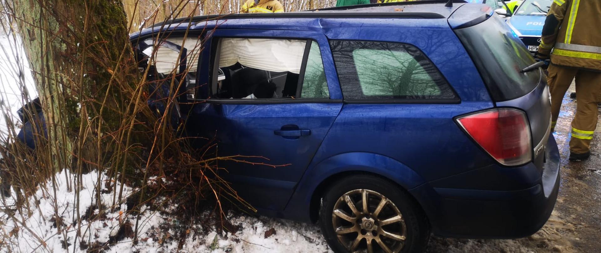 Zdjęcie przedstawia niebieski samochód po zderzeniu z drzewem. W tle strażacy i wóz strażacki