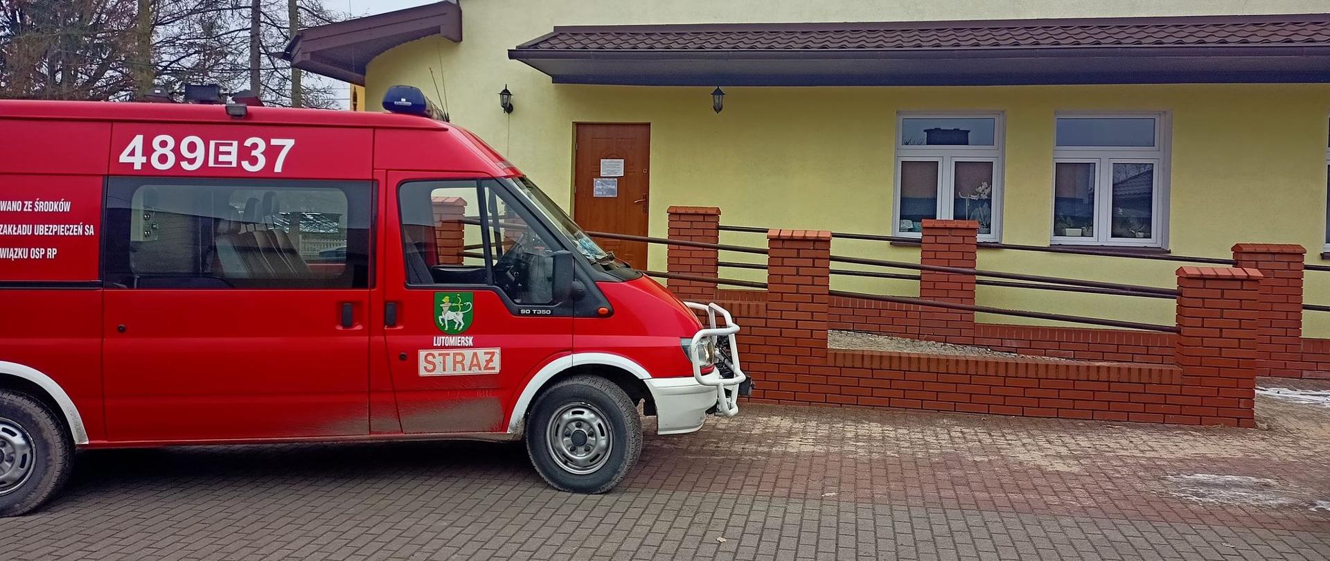 Na pierwszym planie po lewej stronie czerwony pojazd Ochotniczej Straży Pożarnej w Lutomiersku (nr operacyjny 489E37). Na przednich drzwiach pojazdu w białym prostokącie czerwony napis STRAŻ. Powyżej naklejka z herbem gminy Lutomiersk, na zielonej tarczy kroczący centaur strzelający z łuku o barwie żółtej strzałą o barwie żółtej za siebie w kierunku ogona, zakończonego łbem węża o oku barwy żółtej. Na drugim planie budynek punktu szczepień z widocznym podjazdem dla wózków inwalidzkich. Elewacja budynku ma kolor żółty, dach w kolorze brązowym.