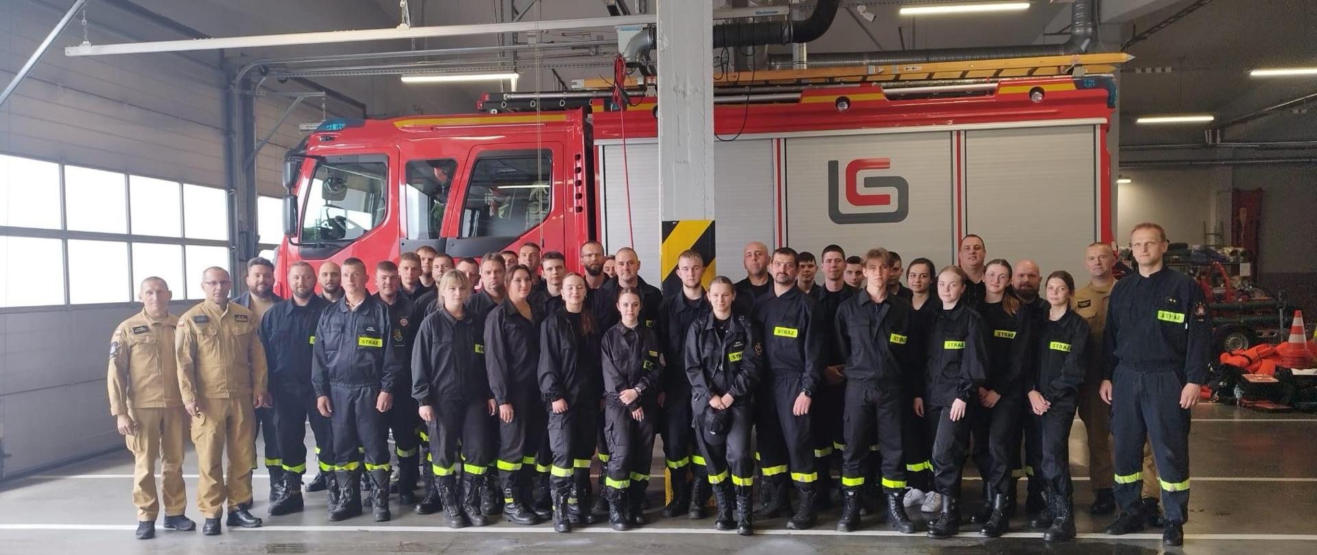 Strażacy i strażaczki stoją w dwuszeregu na garażu na tle pojazdów ratowniczych.