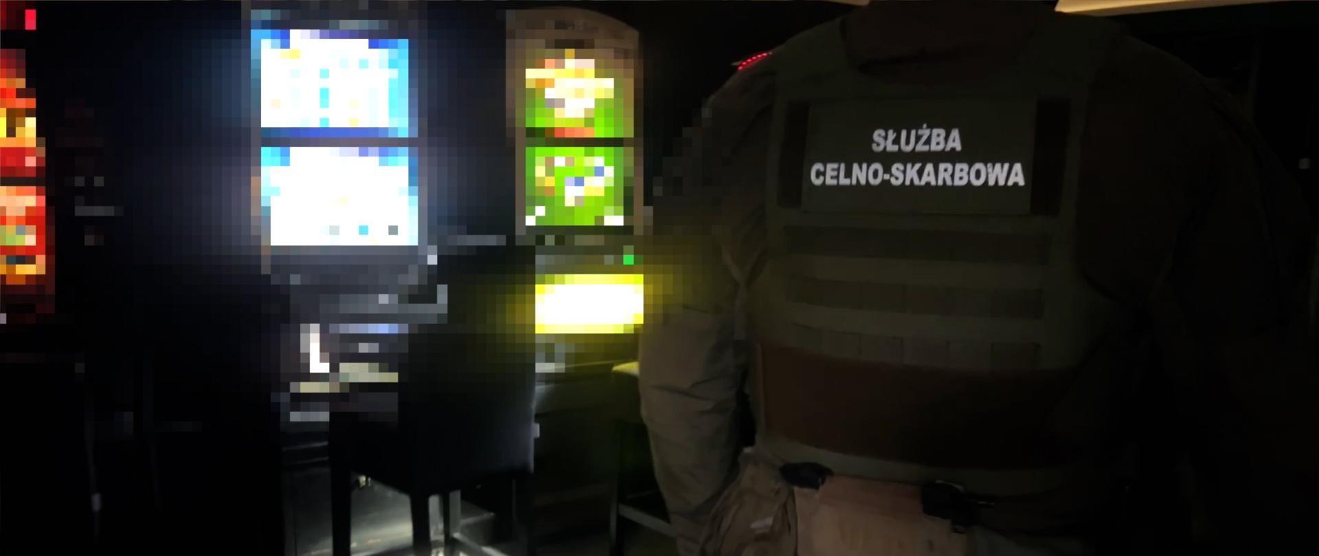 Osoba w kamizelce z napisem „Służba Celno‑Skarbowa” stoi w ciemnym pomieszczeniu obok podświetlonych automatów.