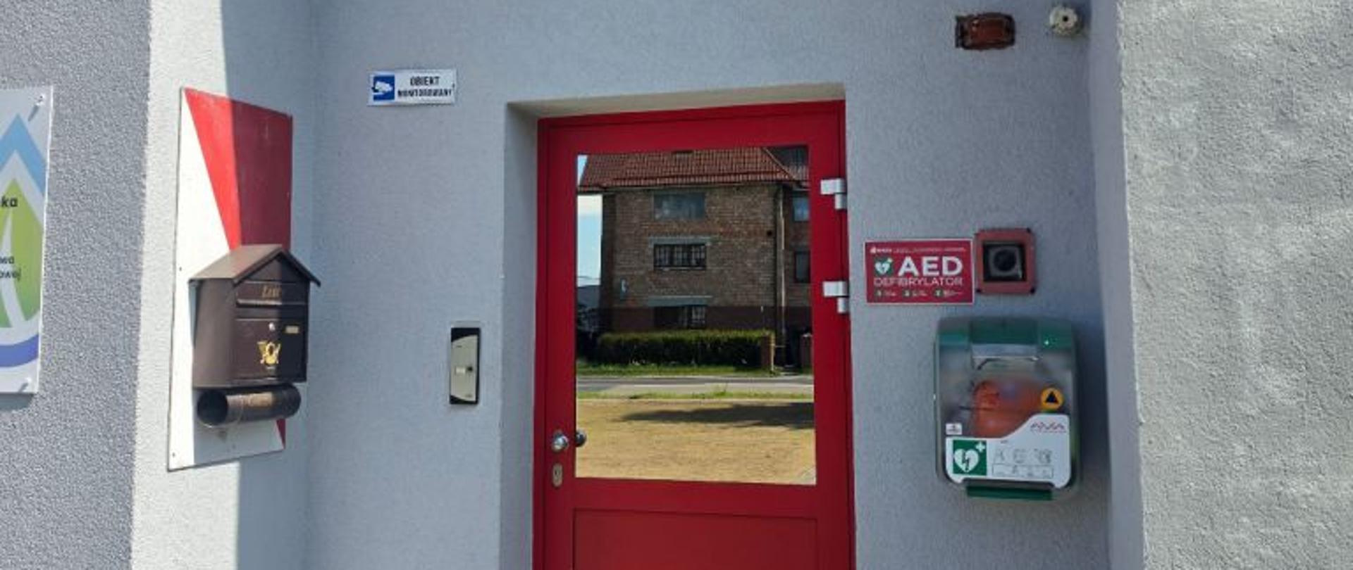 Pionowe zdjęcie przedstawiające główne wejście do budynku wykonanego z jasnoszarego, strukturalnego tynku. W centralnej części kadru znajdują się jednoskrzydłowe, przeszklone drzwi wejściowe w wyrazistej, czerwonej ramie, w których szybie odbija się sąsiedni budynek z cegły oraz bezchmurne niebo. Nad drzwiami zamontowane są dwa elementy identyfikacyjne: owalne godło Polski z Białym Orłem na czerwonym tle, a bezpośrednio pod nim prostokątna, czerwona tablica urzędowa z białym napisem „KOMENDA POWIATOWA PAŃSTWOWEJ STRAŻY POŻARNEJ w Białogardzie woj. zachodniopomorskie”. Po prawej stronie drzwi, na wysokości wzroku, zamontowana jest zielono-szara, plastikowa skrzynka defibrylatora AED marki Aivia z widocznym przez przezroczystą pokrywą pomarańczowym urządzeniem, a nad nią wisi czerwona tabliczka informacyjna z białym napisem „AED DEFIBRYLATOR” oraz czarny włącznik w czerwonej obudowie. Po lewej stronie drzwi widoczna jest brązowa, metalowa skrzynka na listy, fragment biało-czerwonego malowania pasowego na ścianie oraz mała, niebiesko-biała tabliczka z napisem „OBIEKT MONITOROWANY”. Przed wejściem znajduje się podłoże wyłożone szarą kostką brukową, na którą pada wyraźny cień budynku.