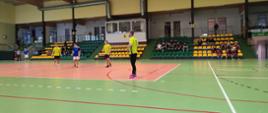 Zawodnicy rywalizujący podczas meczu futsalu w hali sportowej – drużyny w żółtych i niebieskich strojach są w trakcie czekania na wznowienie gry.