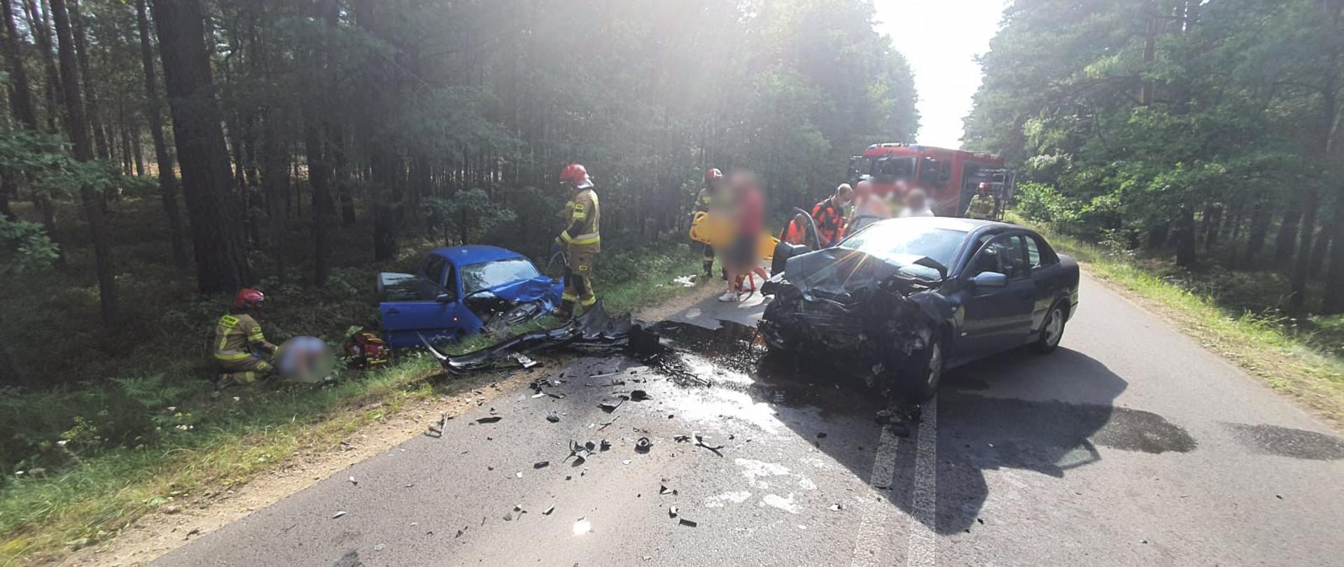Na środku drogi asfaltowej w lesie stoi samochód osobowy z uszkodzonym przodem samochodu. Po lewej stronie w rowie drugi uszkodzony samochód. Przy samochodach strażacy ubrani w ubrania specjalne koloru piaskowego oraz ratownicy medyczni. z tył widoczny samochód pożarniczy.