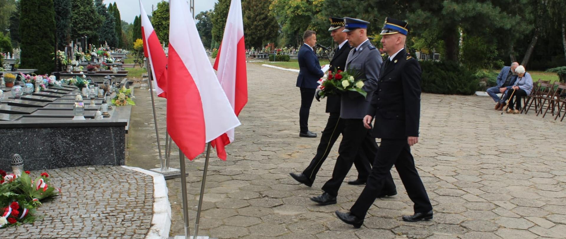 Delegacja służb mundurowych maszeruje złożyć kwiaty pod pomnikiem. Z lewej strony zdjęcia widoczne pomniki grobowe oraz stojaki z flagami RP. Delegacja składa się z 3 formacji: Państwowa Straż Pożarna - funkcjonariusz w umundurowaniu wyjściowym, Policja - funkcjonariusz w umundurowaniu wyjściowym niosący kwiaty, Straż Miejska - funkcjonariusz w umundurowaniu wyjściowym. 