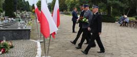 Delegacja służb mundurowych maszeruje złożyć kwiaty pod pomnikiem. Z lewej strony zdjęcia widoczne pomniki grobowe oraz stojaki z flagami RP. Delegacja składa się z 3 formacji: Państwowa Straż Pożarna - funkcjonariusz w umundurowaniu wyjściowym, Policja - funkcjonariusz w umundurowaniu wyjściowym niosący kwiaty, Straż Miejska - funkcjonariusz w umundurowaniu wyjściowym. 