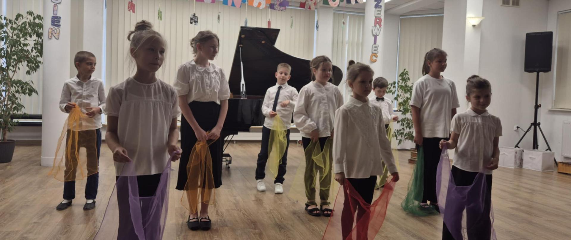 Koncert. W sali stoją uczniowie szkoły muzycznej. Dzieci ubrane są w białe koszule i czarne spodnie lub spódnice. W rękach trzymają kolorowe chusty.