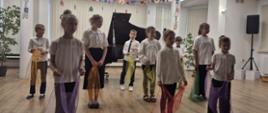 Koncert. W sali stoją uczniowie szkoły muzycznej. Dzieci ubrane są w białe koszule i czarne spodnie lub spódnice. W rękach trzymają kolorowe chusty.