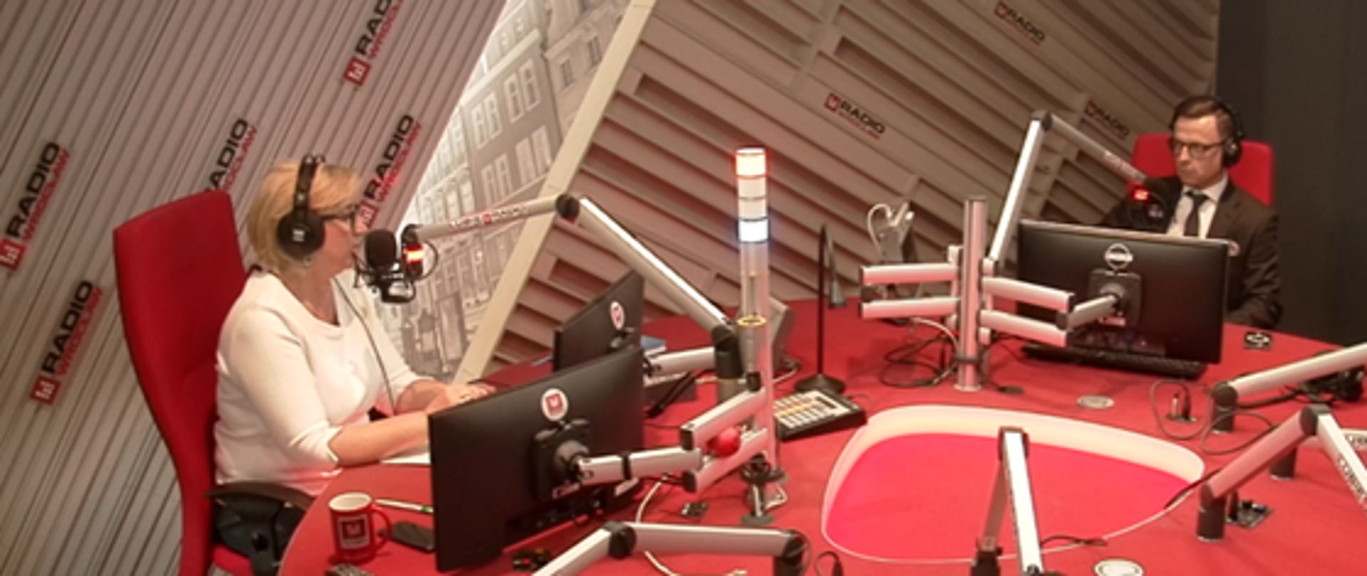 Studio nagraniowe. W centralnej części duży, czerwony stół, na stole ekrany i srebrne ramiona mikrofonowe. 