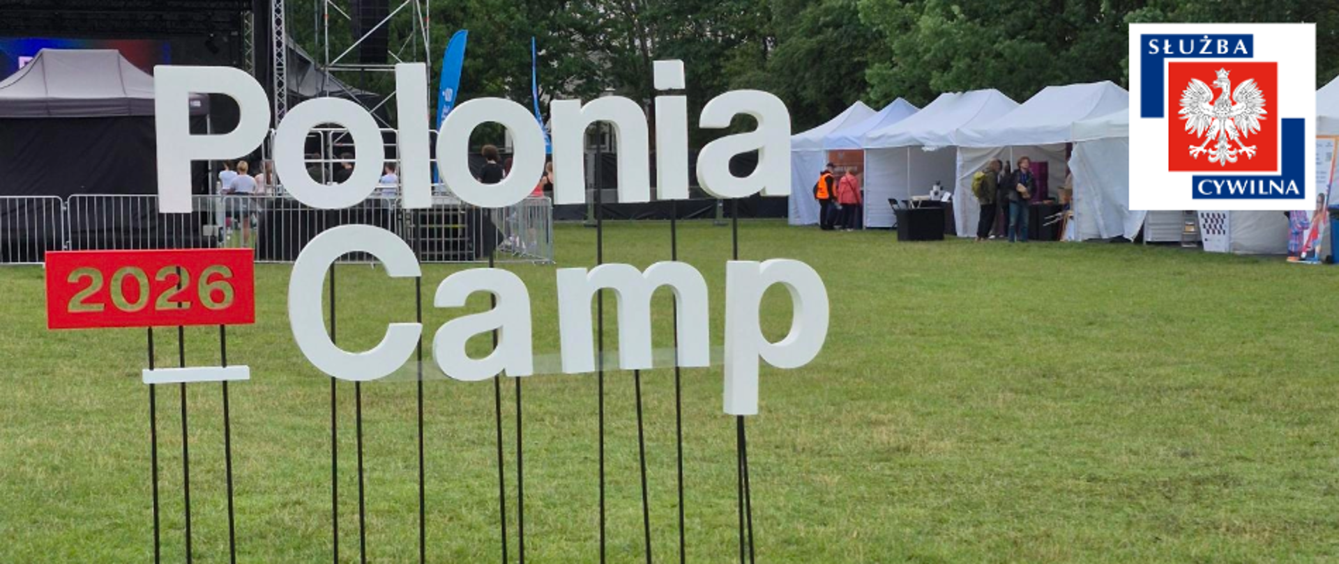 Duży napis „Polonia Camp 2026” na trawniku podczas wydarzenia plenerowego; w tle białe namioty i scena, w prawym górnym rogu logo Służby Cywilnej.