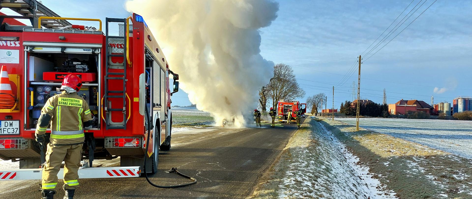 Strażacy Państwowej Straży Pożarnej prowadzą działania gaśnicze przy pożarze samochodu osobowego na drodze lokalnej. Na zdjęciu widoczne są dwa samochody ratowniczo-gaśnicze oraz strażacy zabezpieczający miejsce zdarzenia. Pojazd objęty pożarem znajduje się w gęstej chmurze dymu, która unosi się wysoko nad jezdnią. Akcja prowadzona jest w warunkach zimowych, przy częściowo ośnieżonych poboczach i polach.