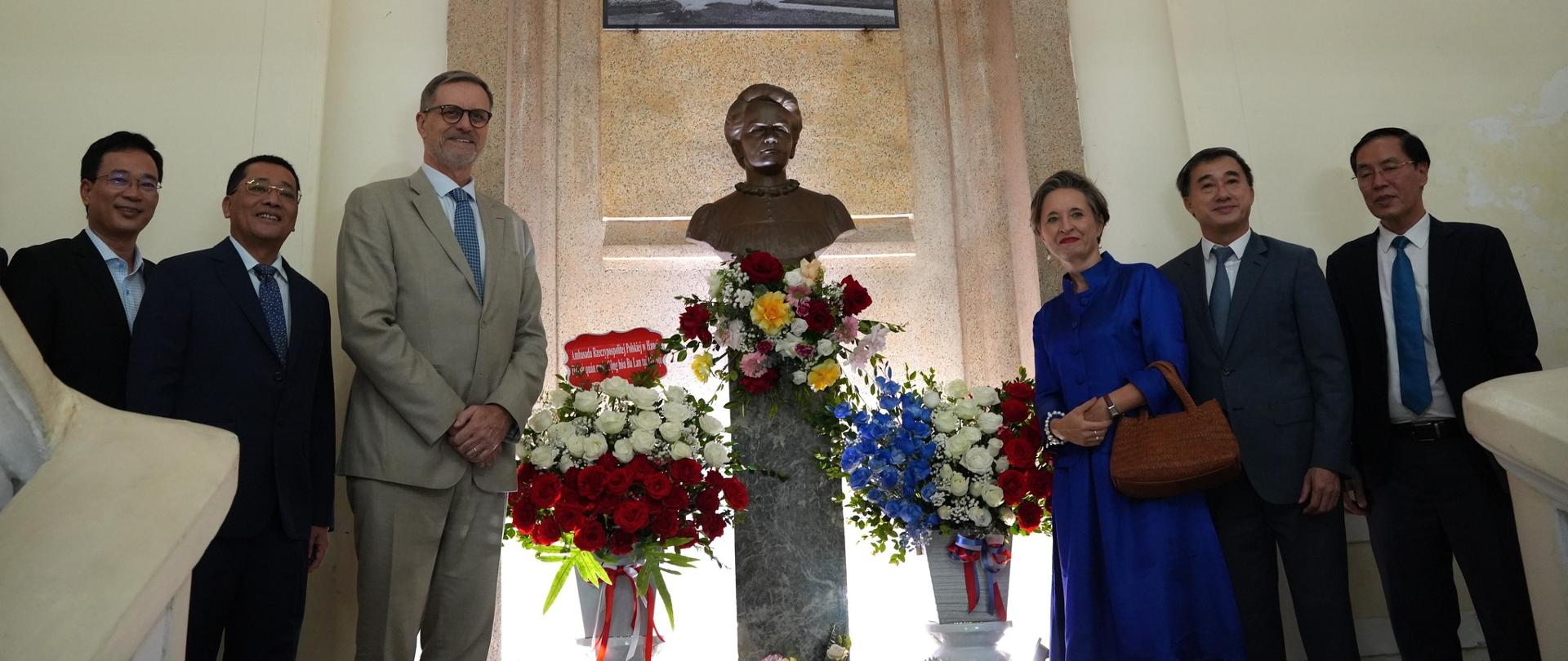 Grupa sześciu osób, w tym kierownicy placówek Francji i Polski - stoi w jasnym wnętrzu przy popiersiu Marii Skłodowskiej-Curie, ustawionym na postumencie z kwiatami; nad nim widoczna tablica pamiątkowa i archiwalne zdjęcie budynku.