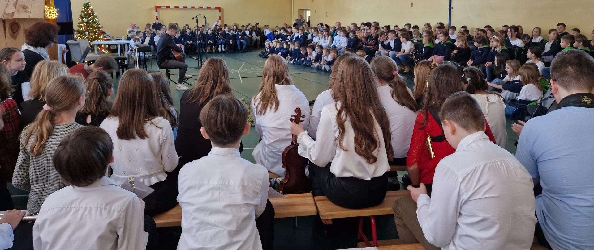 Zdjęcie zostało wykonane na sali gimnastycznej szkoły podstawowej i przedstawia licznie zgromadzoną publiczność siedzącą dookoła oraz wykonawców - uczniów szkoły muzycznej oczekujących na występ. Na środku widać grającego na gitarze ucznia na tle dekoracji - drewnianej stajenki i ubranej choinki. 