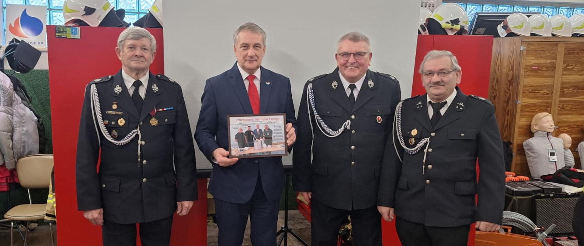 Ministra oraz druhów, którzy stoją obok siebie i pozują do zdjęcia. Minister trzyma w ręce zdjęcie oprawione w ramce.