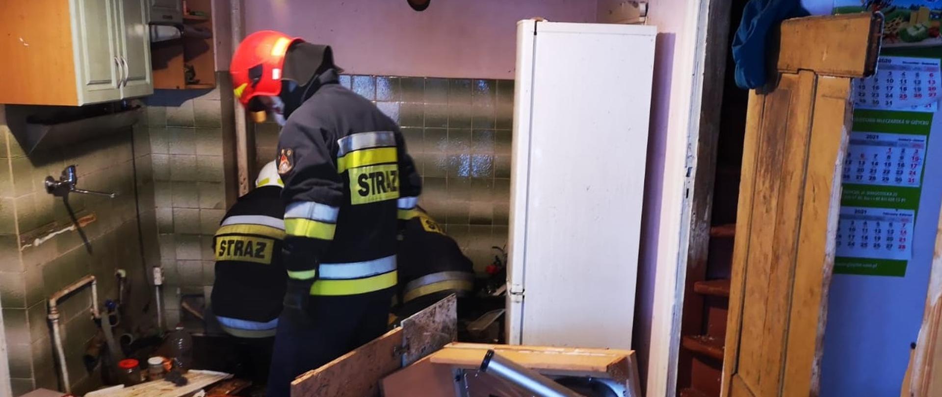 Trzech strażaków w ubraniach specjalnych sprawdzają w zniszczonej kuchni piec c.o, który wybuch. Dookoła znajdują się zniszczone meble. 