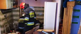 Trzech strażaków w ubraniach specjalnych sprawdzają w zniszczonej kuchni piec c.o, który wybuch. Dookoła znajdują się zniszczone meble. 