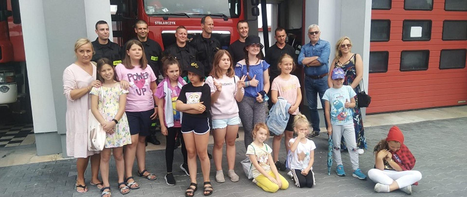 Zdjęcie przedstawia KP PSP w Białogardzie - wizyta dzieci z klubu osiedlowego działającego przy Białogardzkiej Spółdzielni Mieszkaniowej. Akcja ” Bezpieczne Wakacje”. Zdjęcie grupowe dzieci, opiekunów i strażaków przed garażami samochodów strażackich