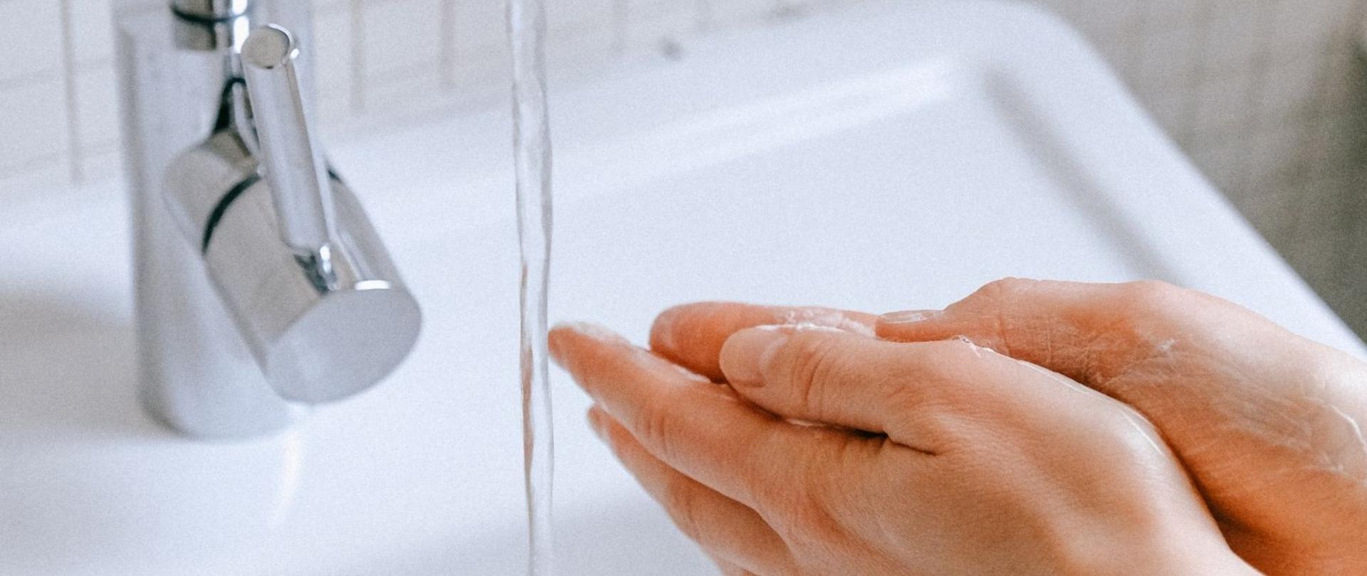 Zdjęcie przedstawia zlew, kran z płynącą wodą, mydło oraz dłonie osoby, która je myje