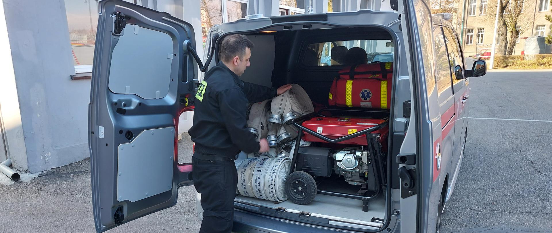 Strażak wkłada wąż pożarniczy do przedziału bagażowego samochodu dostawczego. 