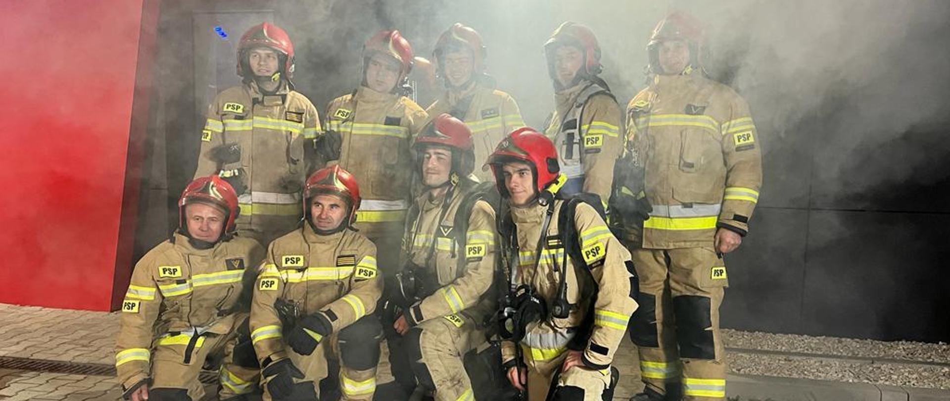 Zdjęcie przedstawia strażaków Jednostki Ratowniczo-Gaśniczej Numer 1 w Kielcach podczas realizacji spotu o bezpieczeństwie na tle zadymionego wyjścia z budynku.