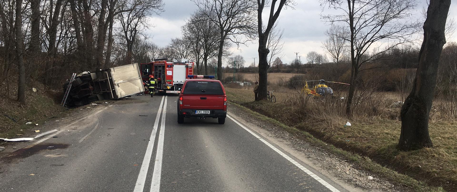 Na zdjeciu widoczy jest jeden samochód dostawczy który lezy na boku w rowie dwa sa,ochody PSP oraz Śmigłowiec LPR na pobliskim polu ronym