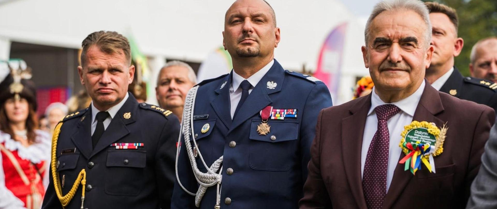 Na pierwszym planie przedstawiciele służb oraz zaproszeni goście uczestniczący w uroczystościach dożynkowych. Wśród gości, pierwszy od lewej, znajduje się Zastępca Kujawsko-Pomorskiego Komendanta Wojewódzkiego PSP st. bryg. Marek Rusoń. Wśród uczestników, w centralnej części, widoczny jest Komendant Powiatowy PSP w Rypinie bryg. Piotr Wypych. W tle pozostali uczestnicy wydarzenia oraz przedstawiciele służb mundurowych.