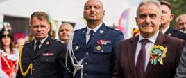 Na pierwszym planie przedstawiciele służb oraz zaproszeni goście uczestniczący w uroczystościach dożynkowych. Wśród gości, pierwszy od lewej, znajduje się Zastępca Kujawsko-Pomorskiego Komendanta Wojewódzkiego PSP st. bryg. Marek Rusoń. Wśród uczestników, w centralnej części, widoczny jest Komendant Powiatowy PSP w Rypinie bryg. Piotr Wypych. W tle pozostali uczestnicy wydarzenia oraz przedstawiciele służb mundurowych.