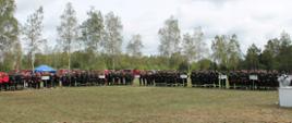 Na fotografii na płycie stadionu stoją w szeregu członkowie drużyn jednostek Ochotniczych Straży Pożarnej, które biorą udział w zawodach. Są ustawieni w grupach podzielonych na poszczególne gminy. Pierwszy zawodnik w grupie trzyma tablicę z nazwą gminy jaką zespoły reprezentują. Zawodnicy ubrani w umundurowanie polowe oraz dresy sportowe wymagane w zawodach. Na głowach zawodnicy mają ubrane hełmy strażackie. Zdjęcie wykonane podczas zbiórki przed zawodami. 