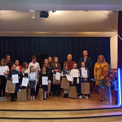 Zdjęcie przedstawia uczniów stojących na scenie z prezentami oraz uniesionymi dyplomami. Po prawej stronie jest trzech nauczycieli. 