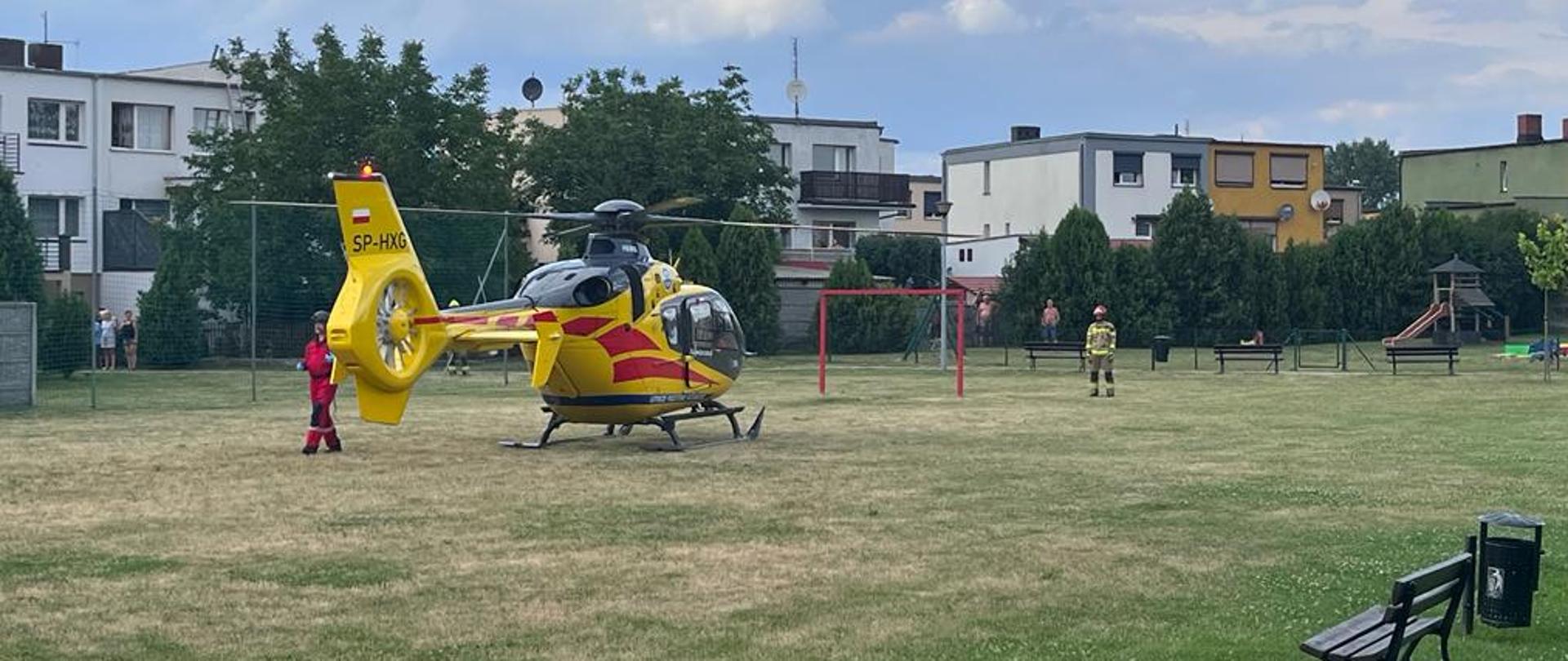 Na zdjęciu helikopter na boisku ławki bramki i strażak obok helikoptera ratownik medyczny za boiskiem budynki mieszkalne