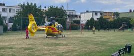 Na zdjęciu helikopter na boisku ławki bramki i strażak obok helikoptera ratownik medyczny za boiskiem budynki mieszkalne