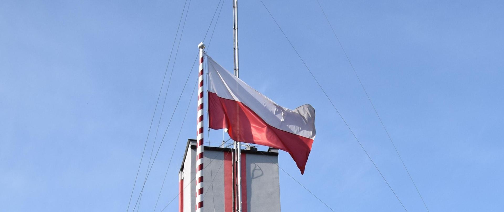 Poczet flagowy wznosi flagę państwową na maszt przed budynkiem KP PSP w Mińsku Mazowieckim podczas uroczystej zmiany służby. Obecni są funkcjonariusze ze zmiany zdającej, przyjmującej, Zastępca Dowódcy JRG oraz Komendant Powiatowy PSP
