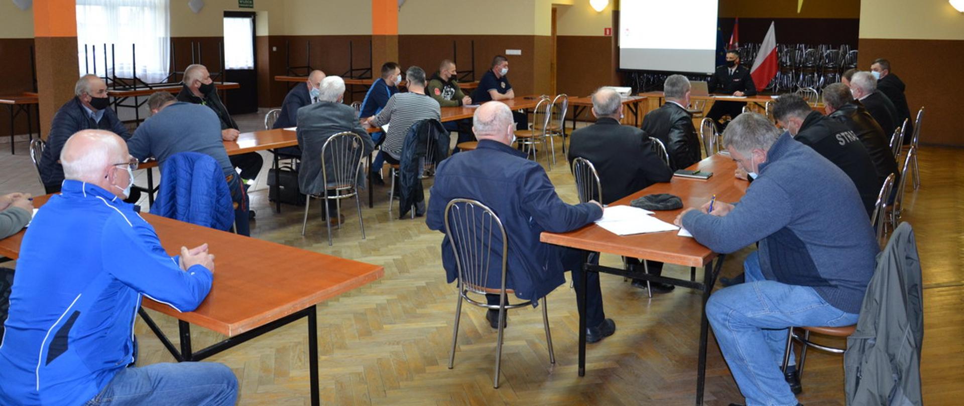 Strażacy siedzą za stołami. przed sceną za stołem siedzi prowadzący spotkanie komendant Powiatowy. Na scenie stoi ekran na którym prezentowane są slajdy.