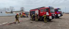 Trzy samochody pożarnicze stoją na betonowym placu. Obok samochodów stoją strażacy w ubraniach specjalnych.